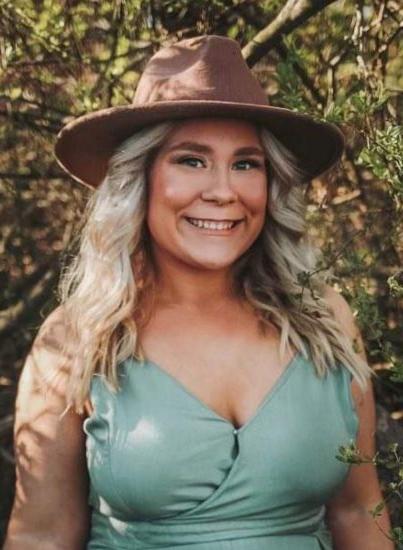 A woman with long blonde hair is smiling outdoors. She is wearing a wide-brimmed brown hat and a light blue sleeveless dress. The background features leafy trees and sunlight filtering through the branches.